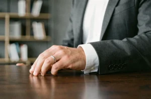 Homme en costume portant une bague au petit doigt gauche posée sur un bureau en bois sombre