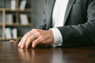Homme en costume portant une bague au petit doigt gauche posée sur un bureau en bois sombre